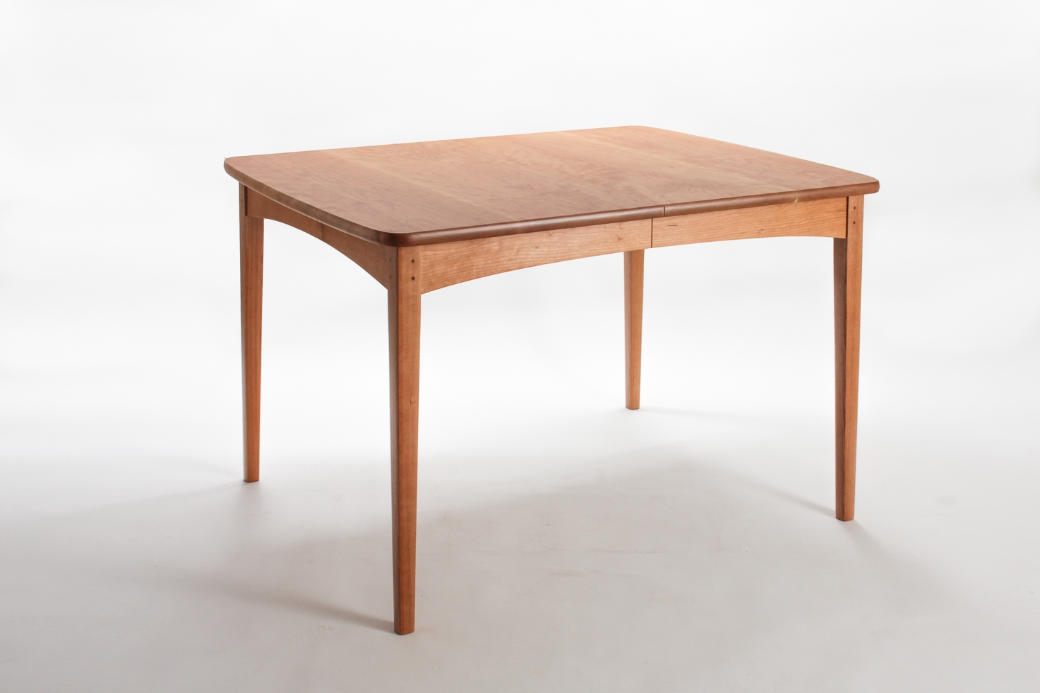 A corner studio view of our cherry Phillips Table. It has tapered, square legs with a thumbnail surface, arched apron, and top that opens for leaves.